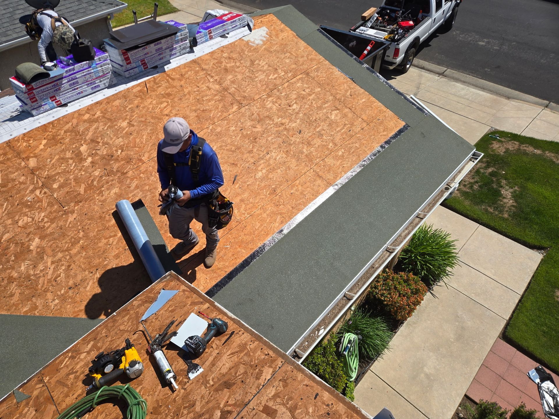Worker on a roof installing roofing materials beside tools and a ladder.