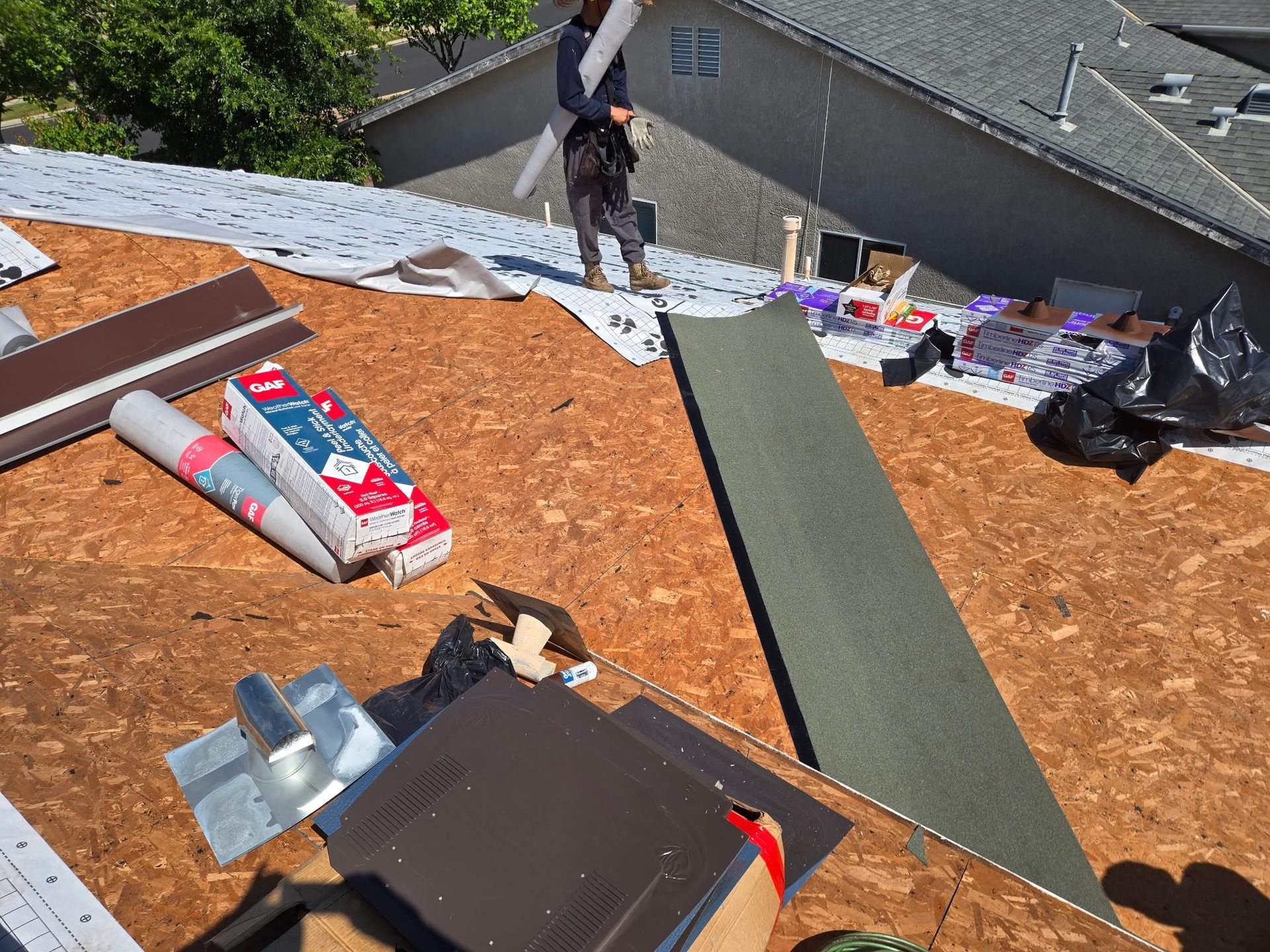 Roofing materials and tools on a rooftop, with a worker standing near the edge below.
