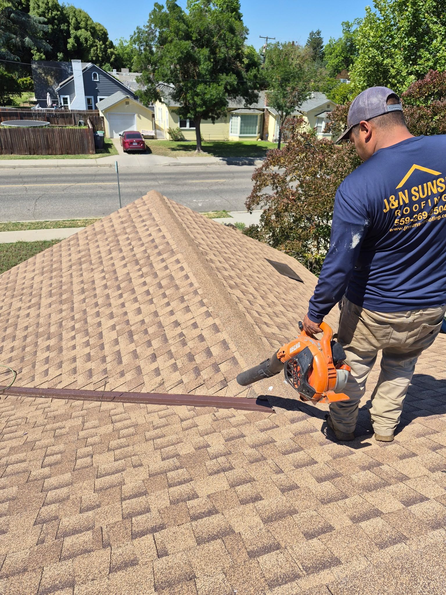 Worker using an orange saw on roof shingles near a house in a suburban neighborhood