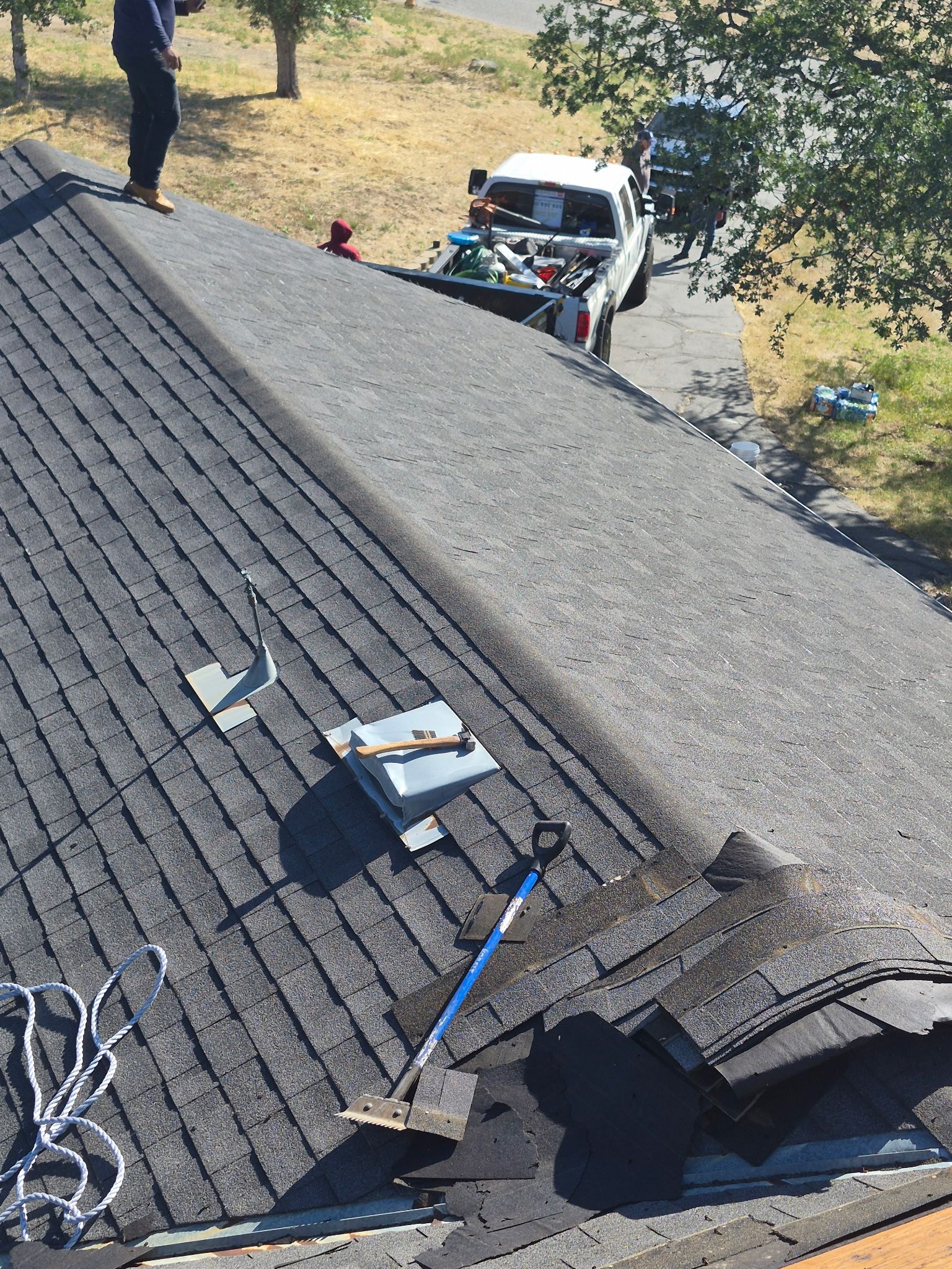 Roof with tools and loose shingles above a driveway where a white work truck is parked.
