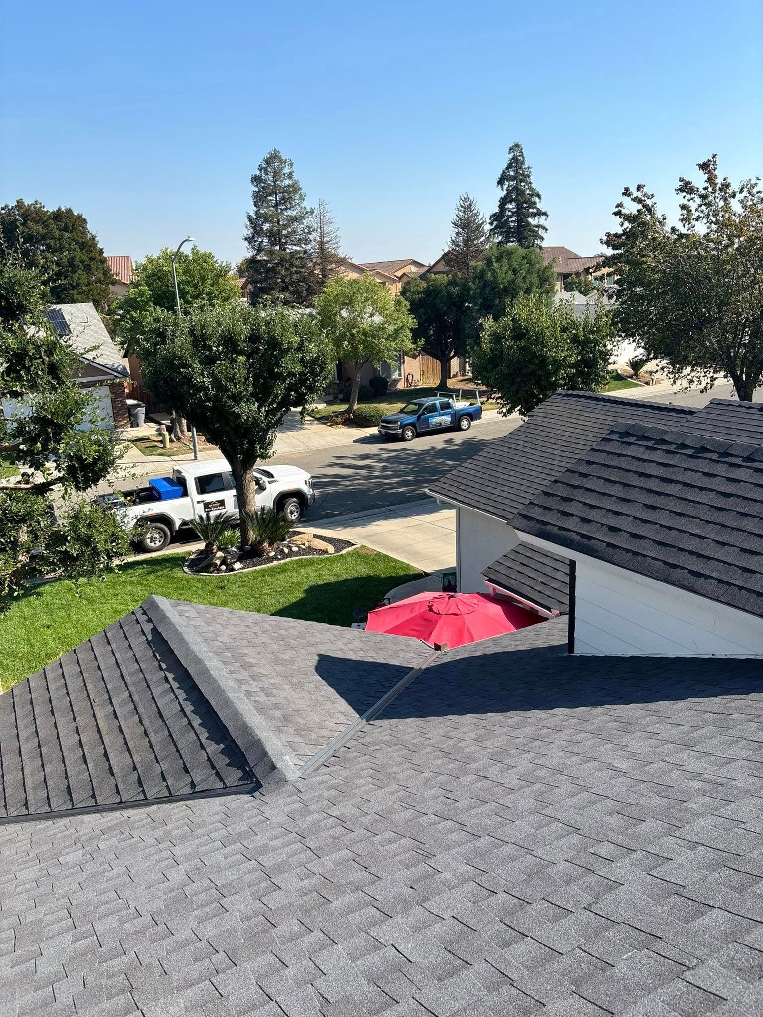 Rooftop view over gray shingles to a sunny suburban street with trees, houses, parked cars, and a red patio umbrella