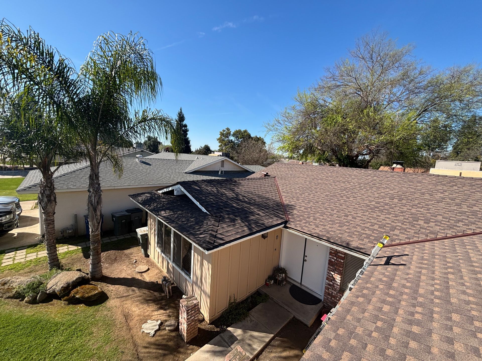 Backyard view of a tan house with brown roof, palm trees, and a sunny blue sky