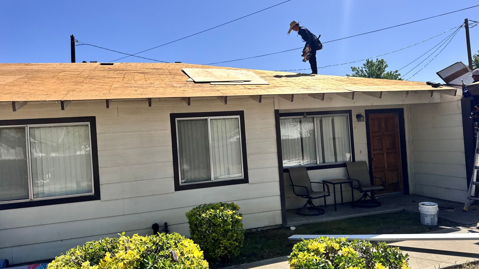 Worker on a roof repairing damaged shingles above a small house with a porch.