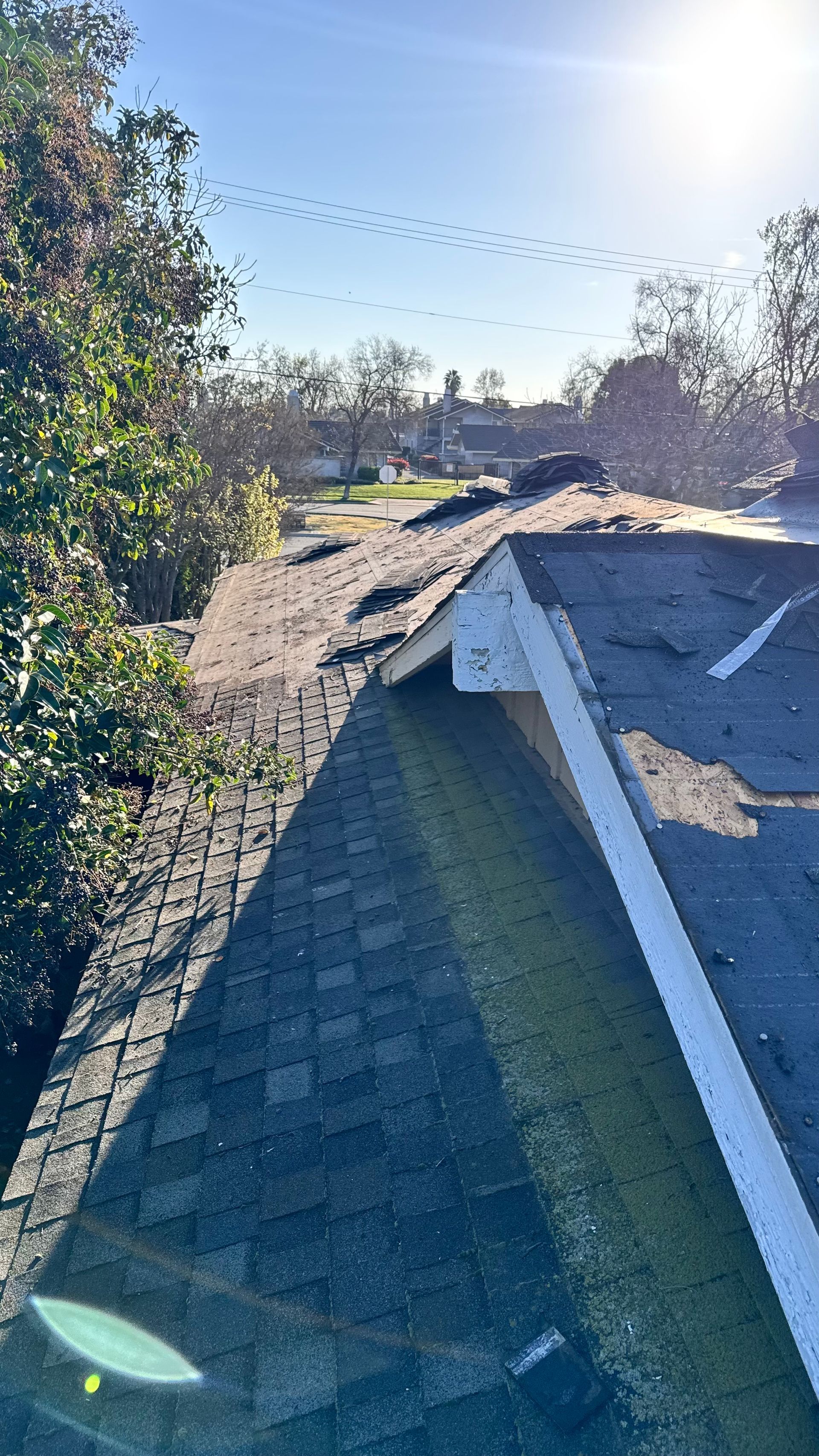 Damaged shingle roof with missing sections and debris in bright sunlight outdoors