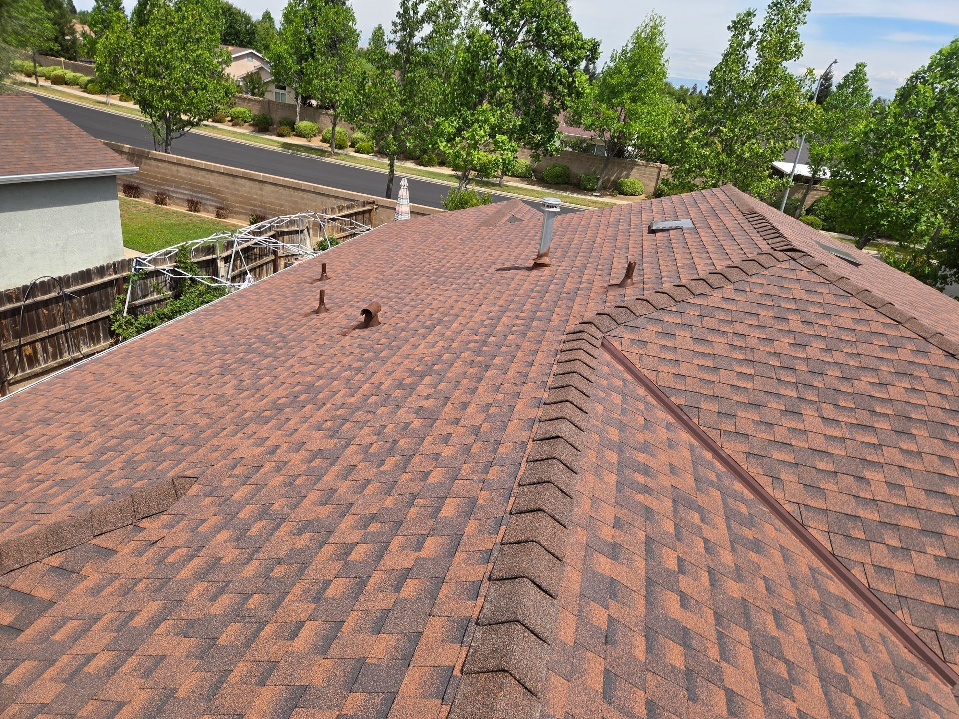 Reddish-brown shingle roof with vents, overlooking trees and a road in the background