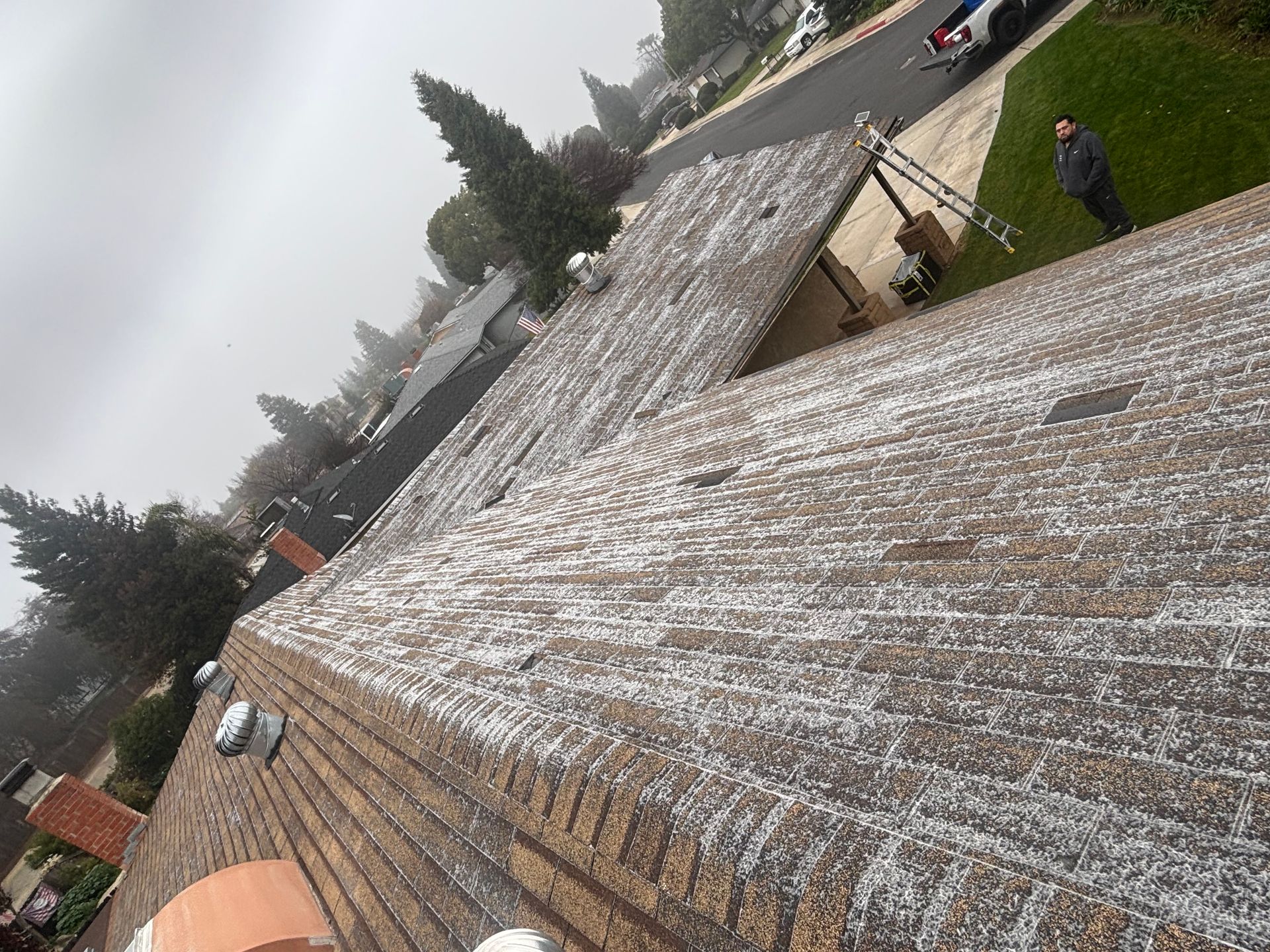 Snow-dusted rooftops in a suburban neighborhood under a cloudy sky, with trees and parked cars nearby