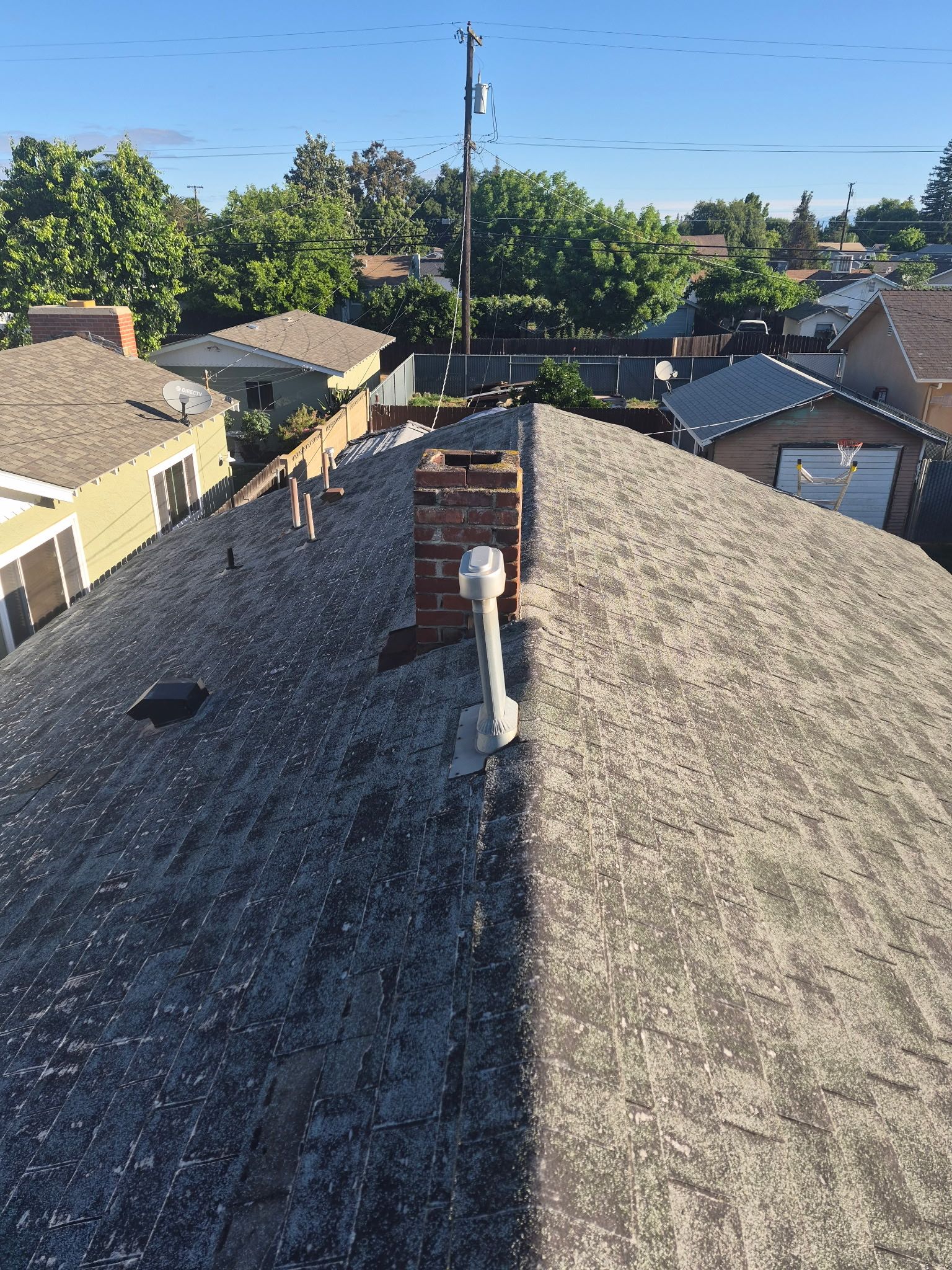 Rooftop view over a neighborhood, showing gray shingles, a vent pipe, and trees under a clear blue sky