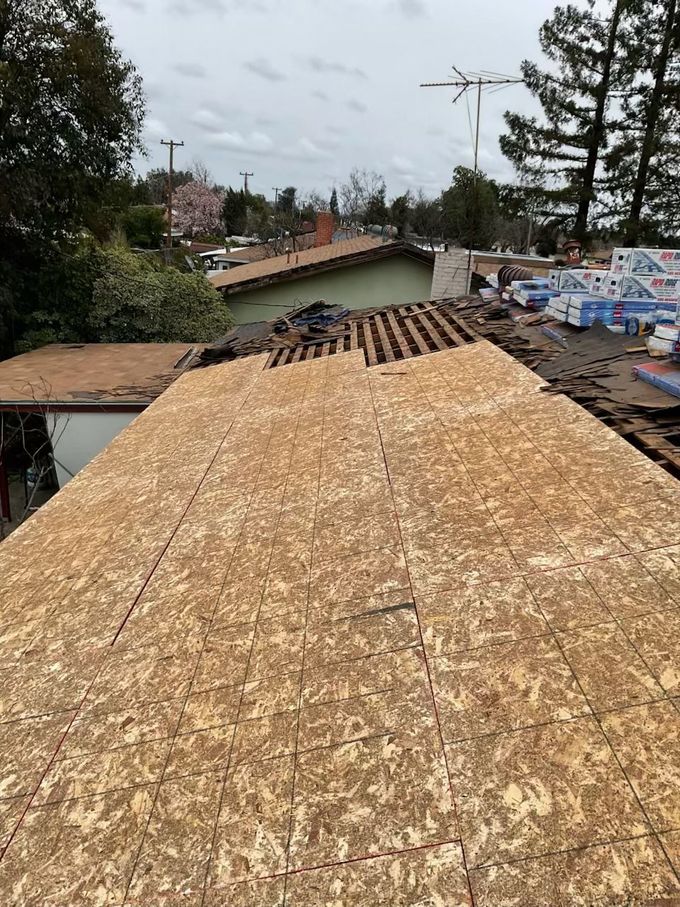 Roof under construction with plywood sheathing, trees, and neighboring houses under a cloudy sky