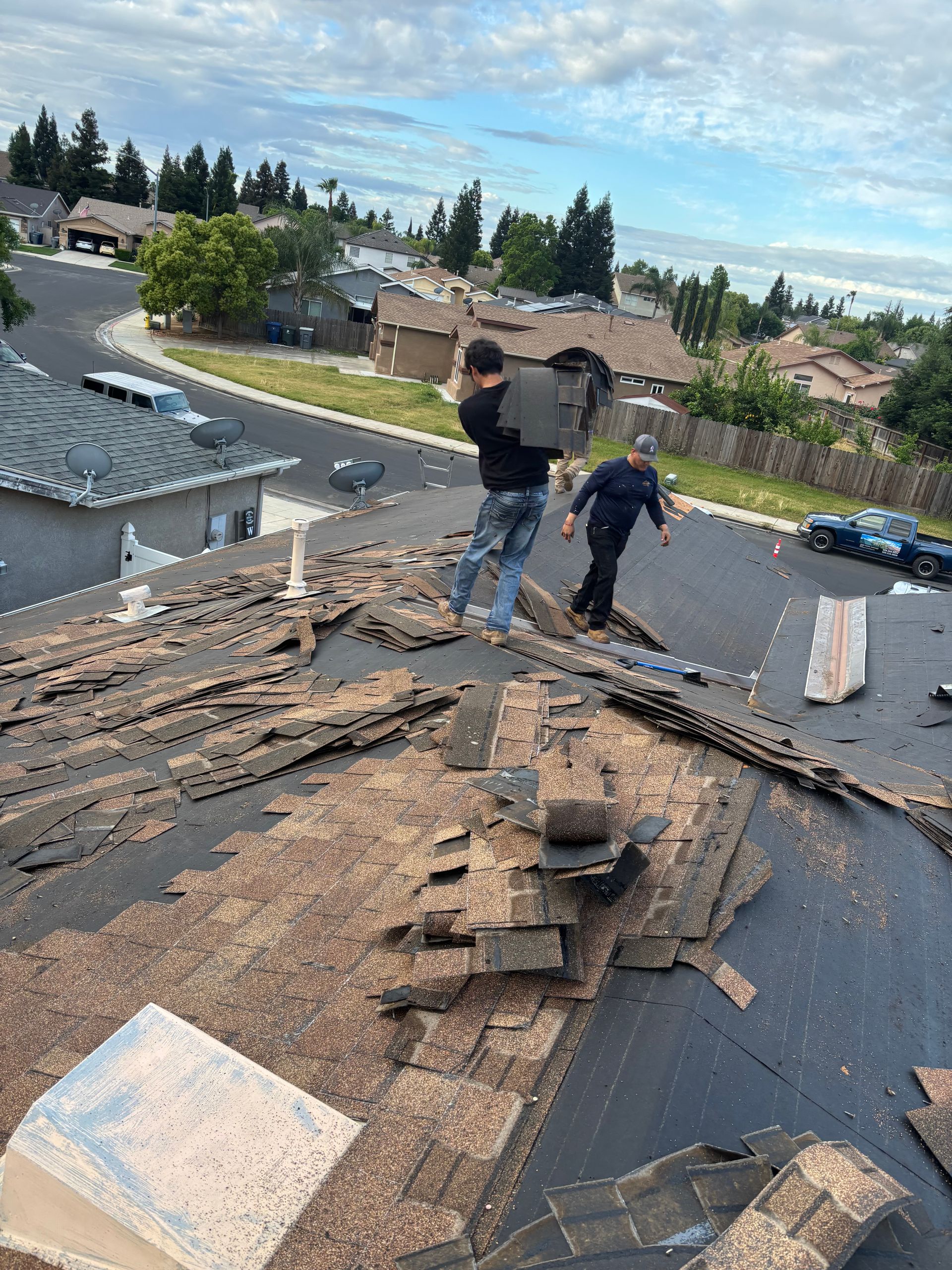 Two workers removing roofing on a house under repair, with shingles and debris spread across the rooftop.