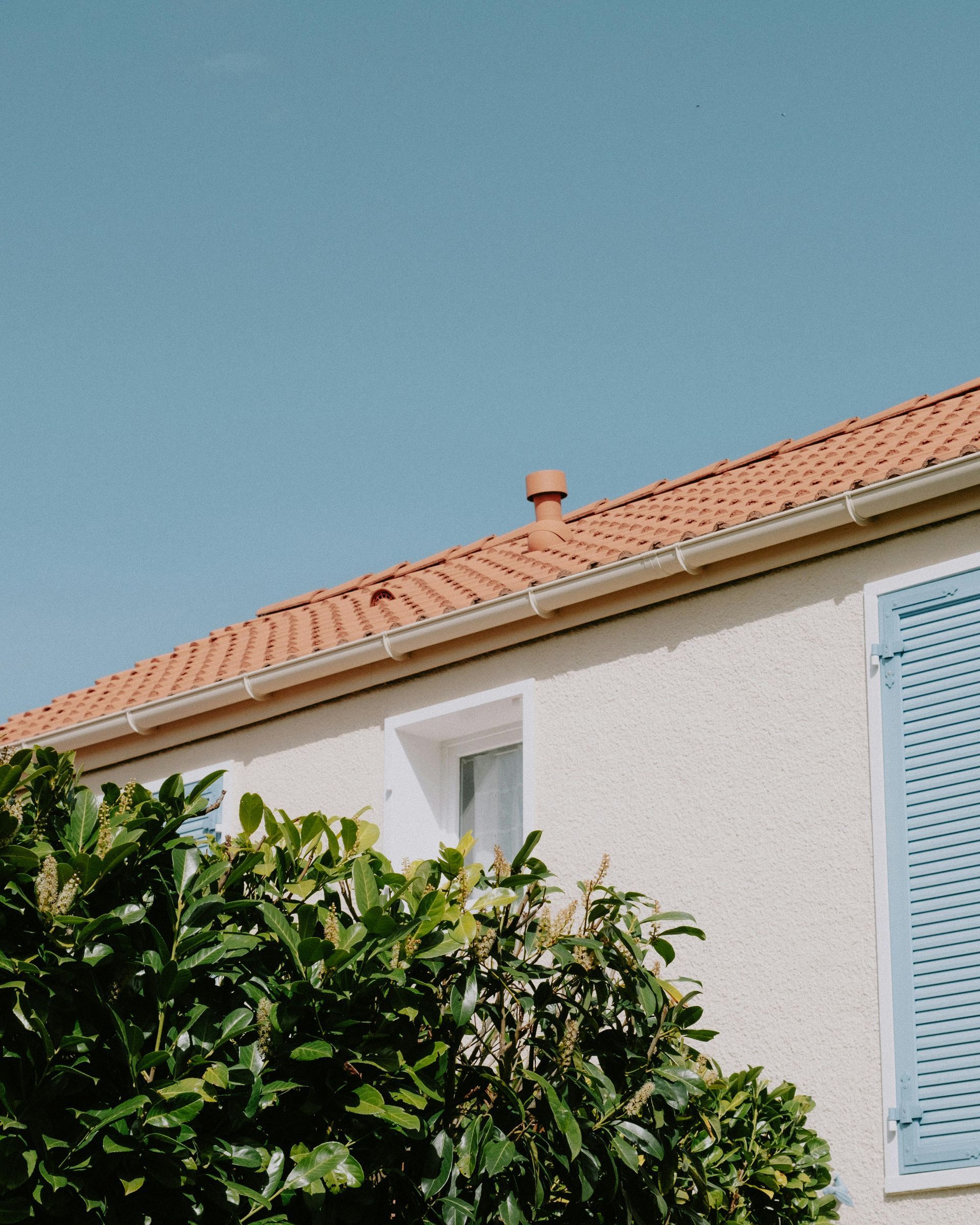 White house with terracotta roof, blue shutters, and leafy green tree against a clear blue sky
