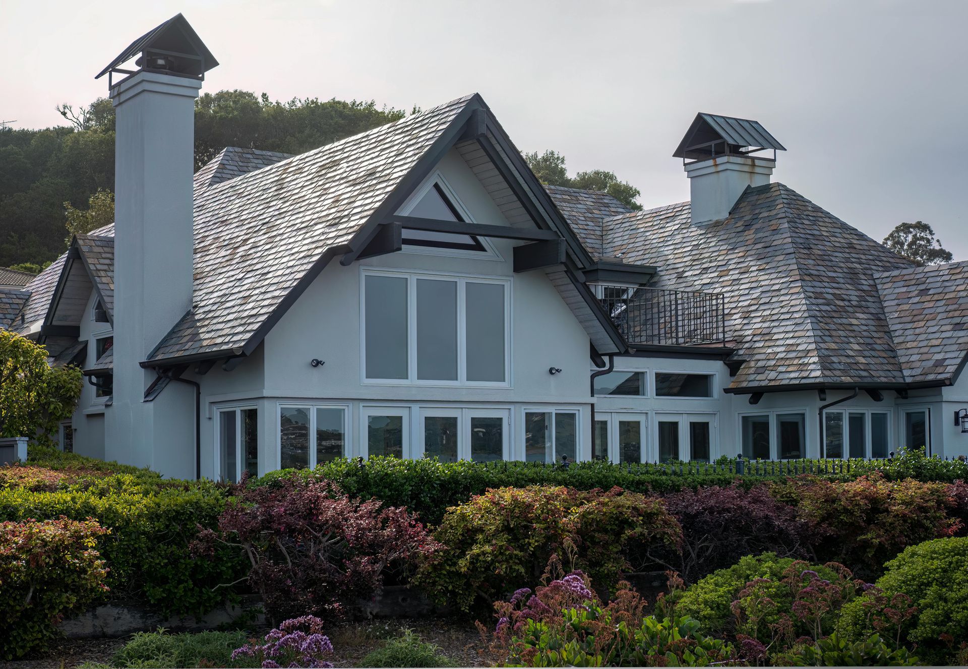 Large white house with a gray shingled roof and chimneys, surrounded by dense green shrubs.
