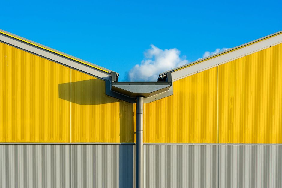 Yellow and gray warehouse roofs meeting at a central gutter under a blue sky with clouds