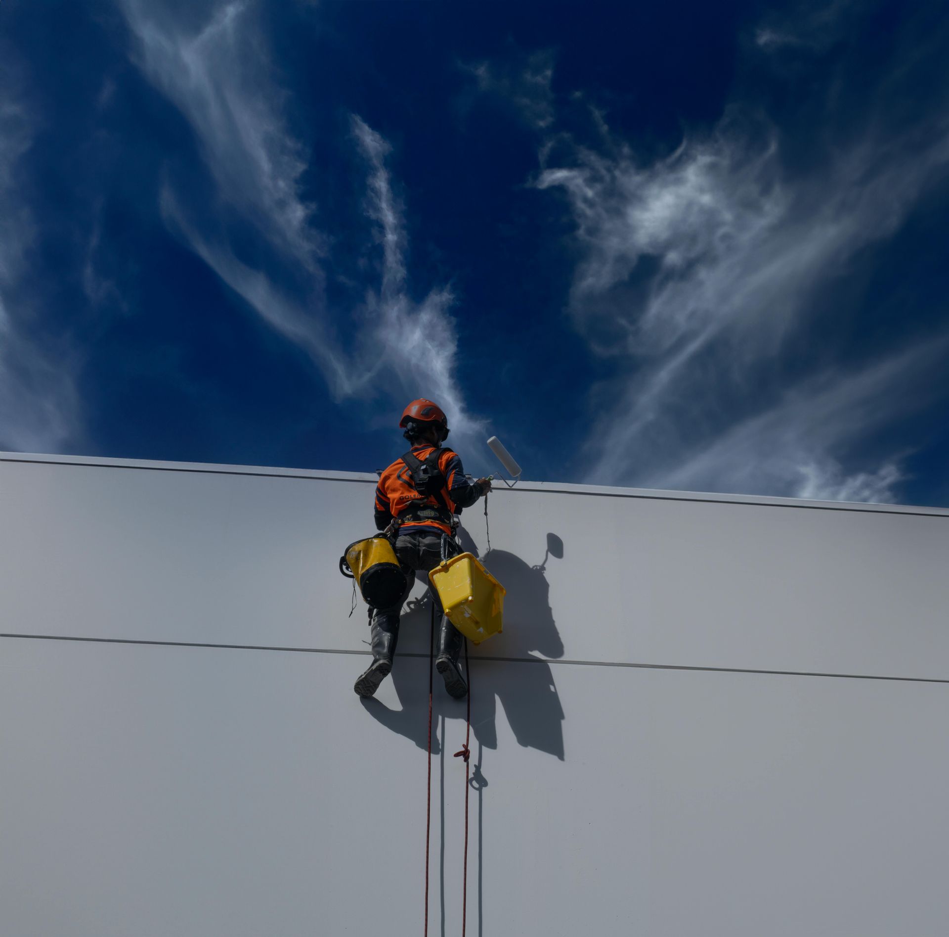 Climber in orange gear on a white wall, hanging by ropes beneath a dramatic blue sky