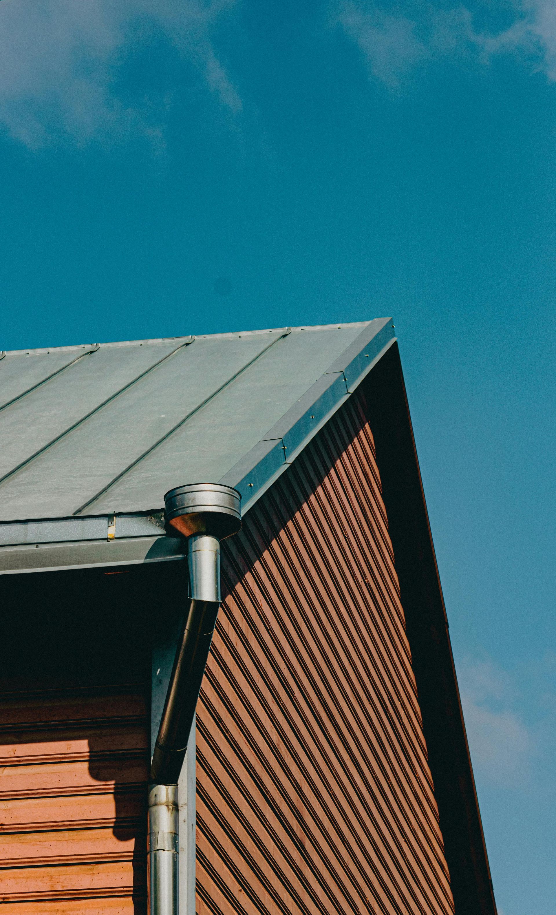 Brick building corner with a gray metal roof and downspout against a blue sky