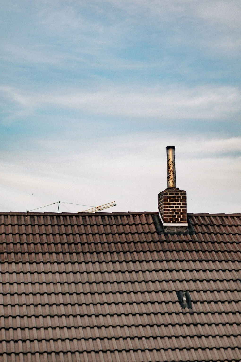 Rooftop with brown tiles and a brick chimney beneath a blue, cloudy sky