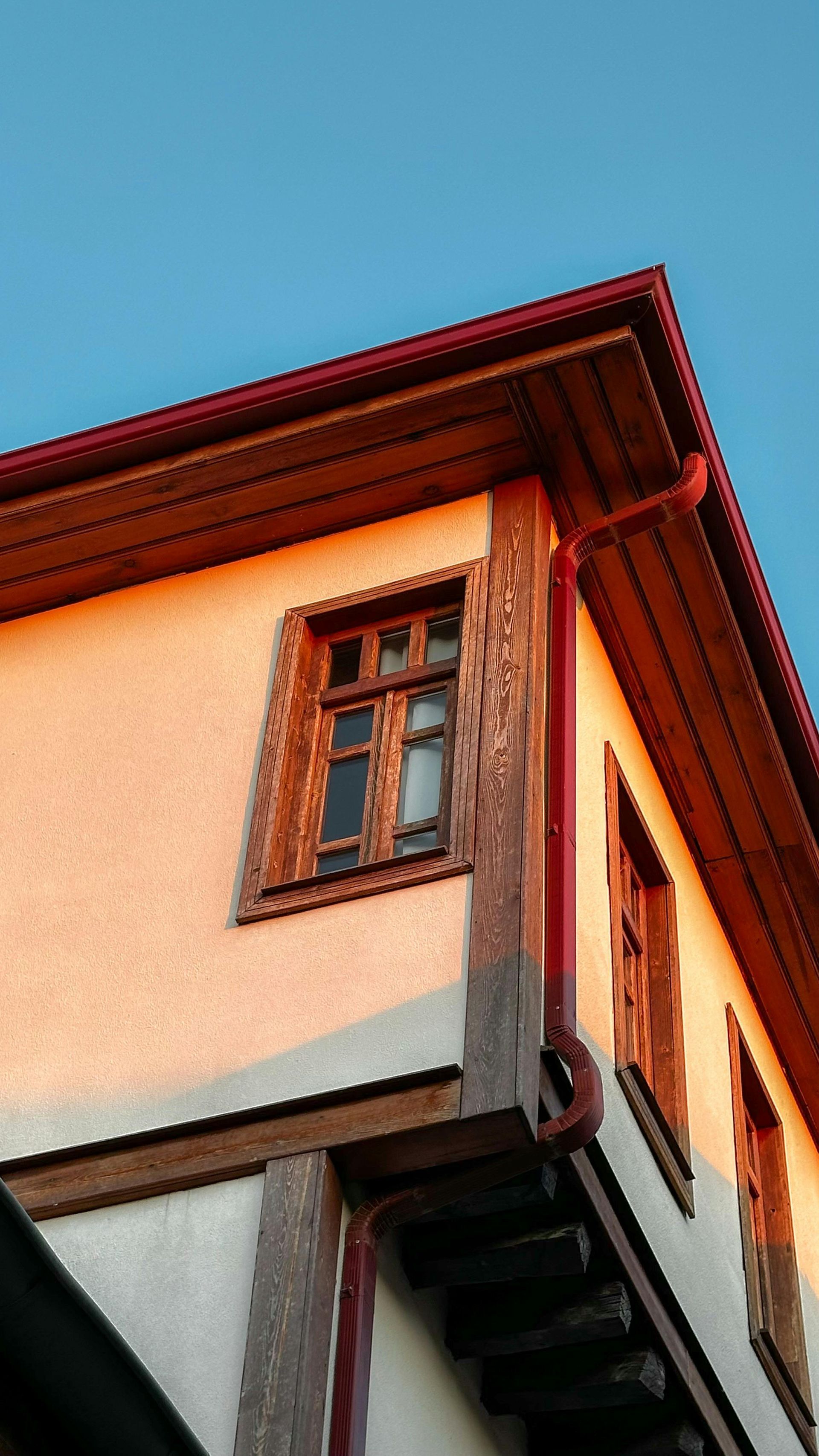 Corner of a cream-and-brown building with a wooden window and red roof trim against blue sky