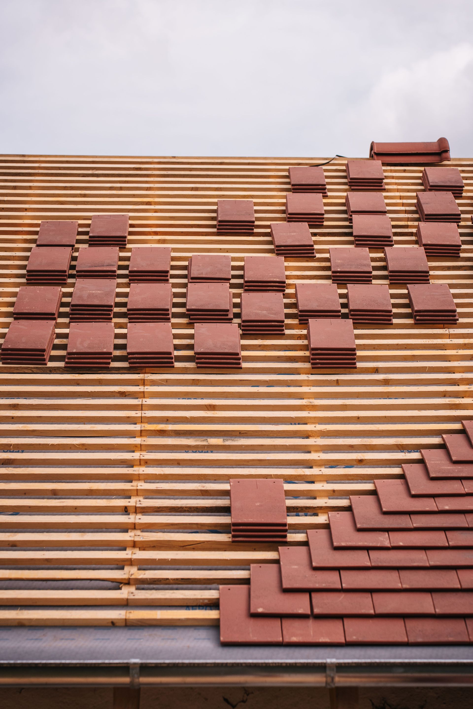 Partially tiled roof with scattered reddish-brown roof tiles on wooden slats under a cloudy sky
