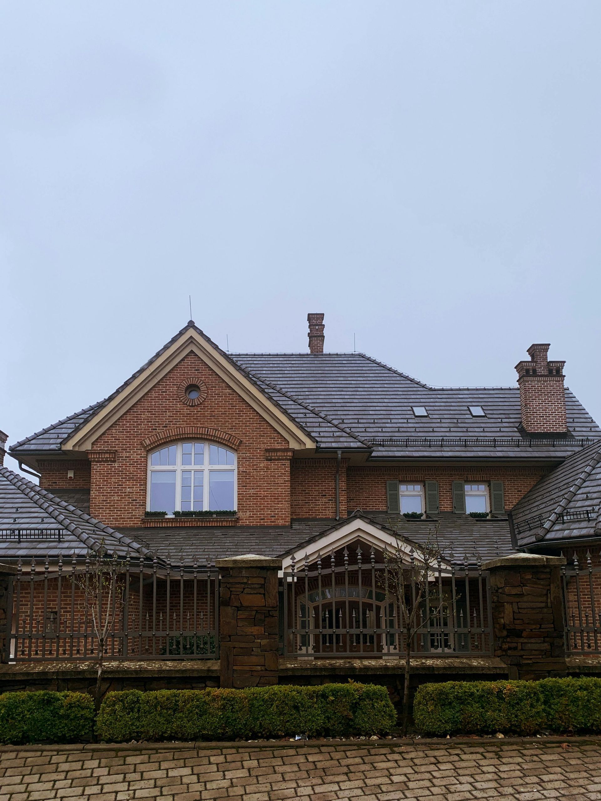 Brick house with dark gray roof, dormer windows, and a front hedge under an overcast sky