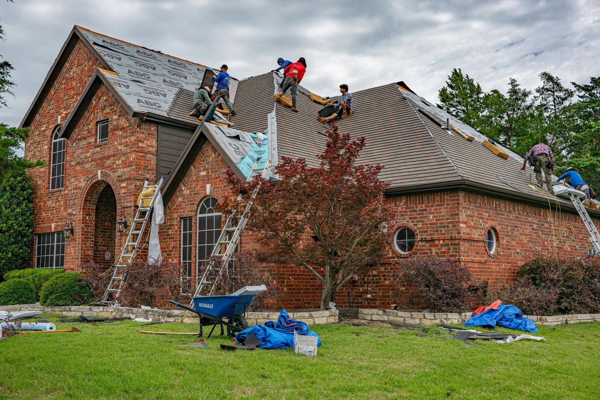 Workers repairing a brick house roof with blue tarps and ladders in a grassy yard