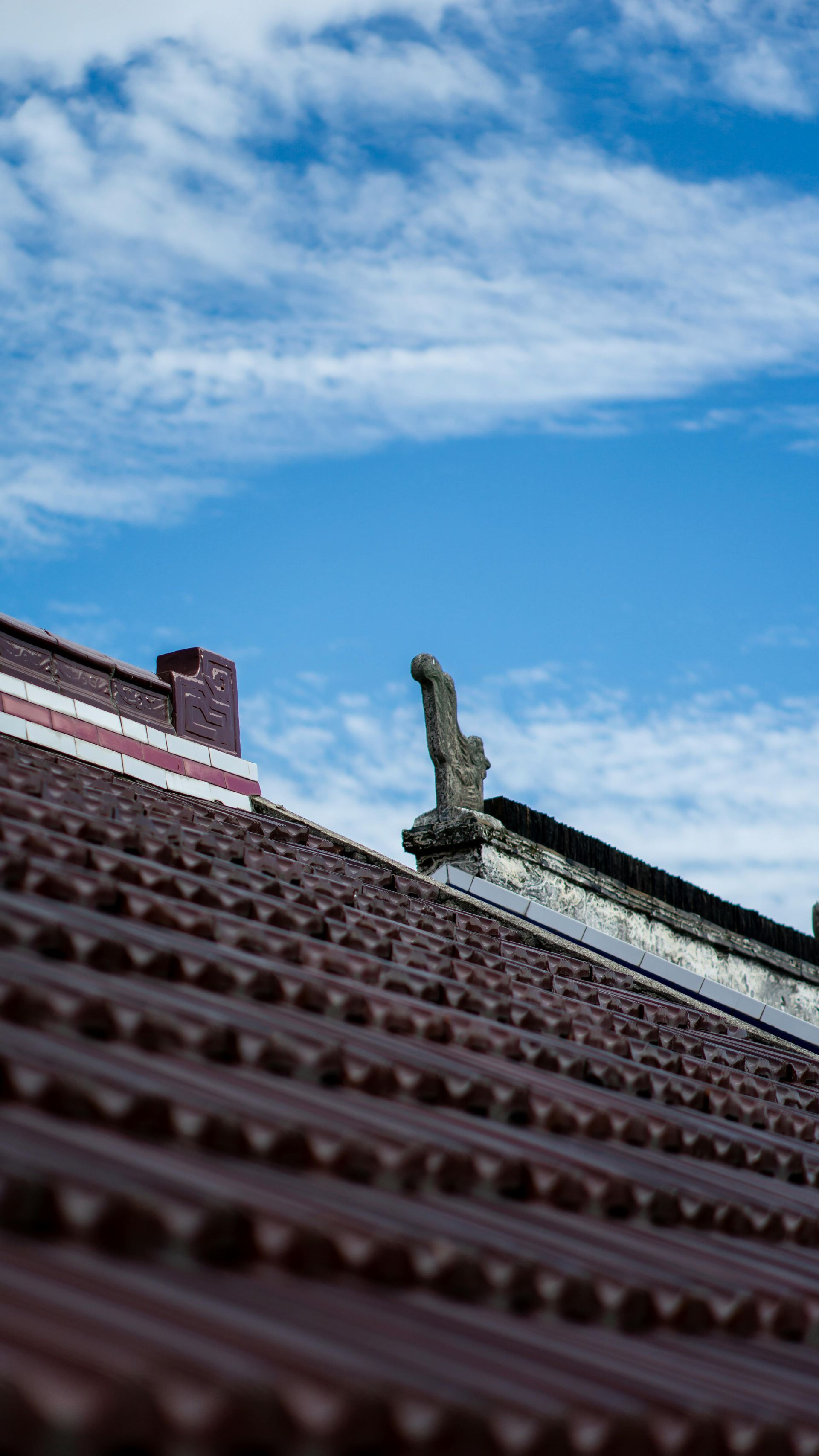Low-angle view of a tiled roof with a statue against a bright blue, cloudy sky