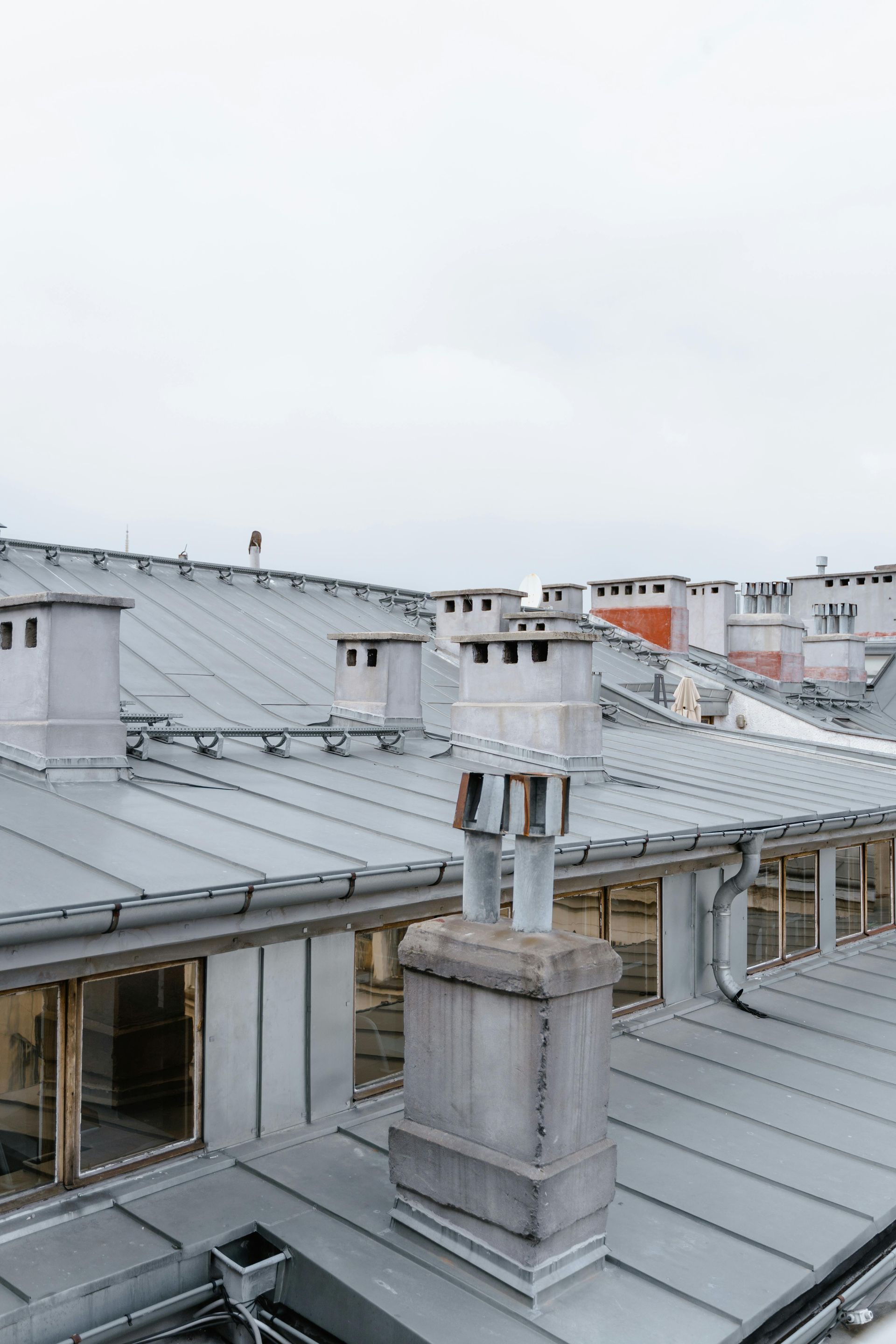 Rooftop with gray metal roofs and chimneys under a pale overcast sky