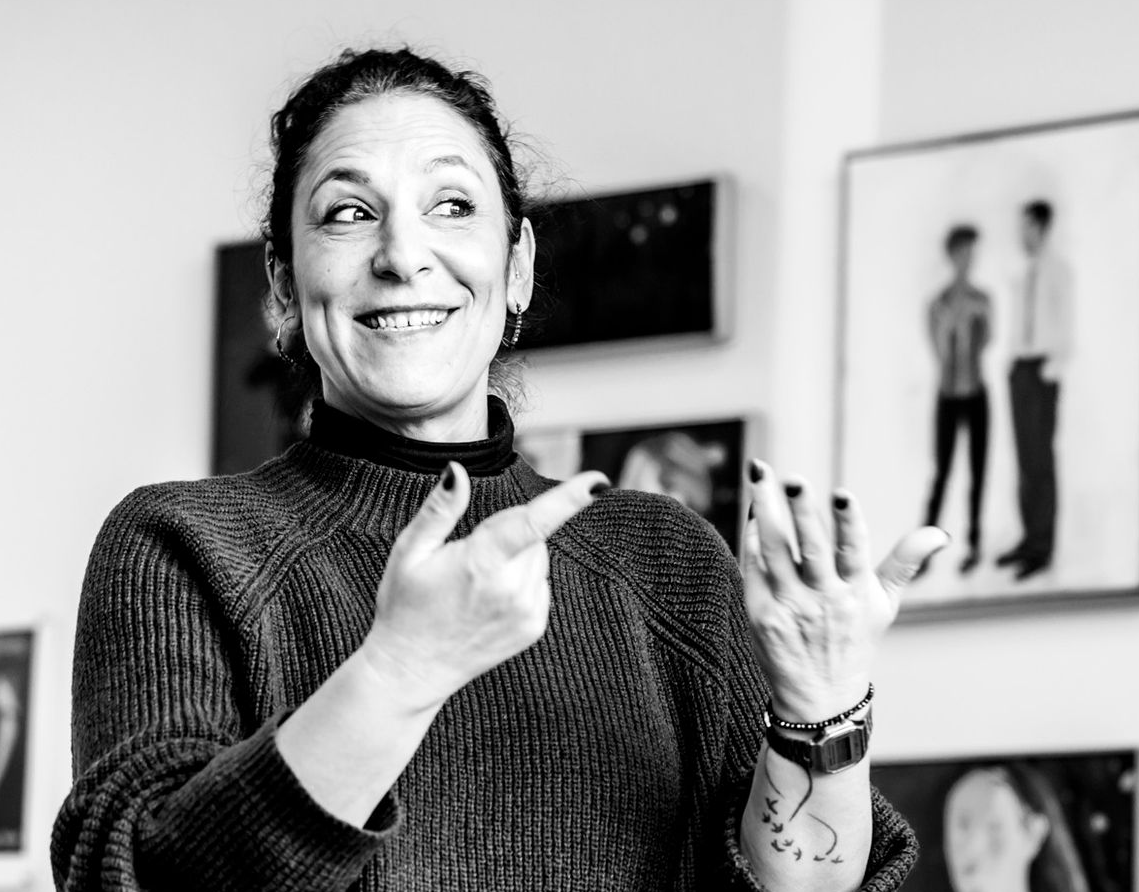 Frau in dunklem Pullover zeigt mit einer Hand und gestikuliert mit der anderen; lächelnd in einer Kunstgalerie.