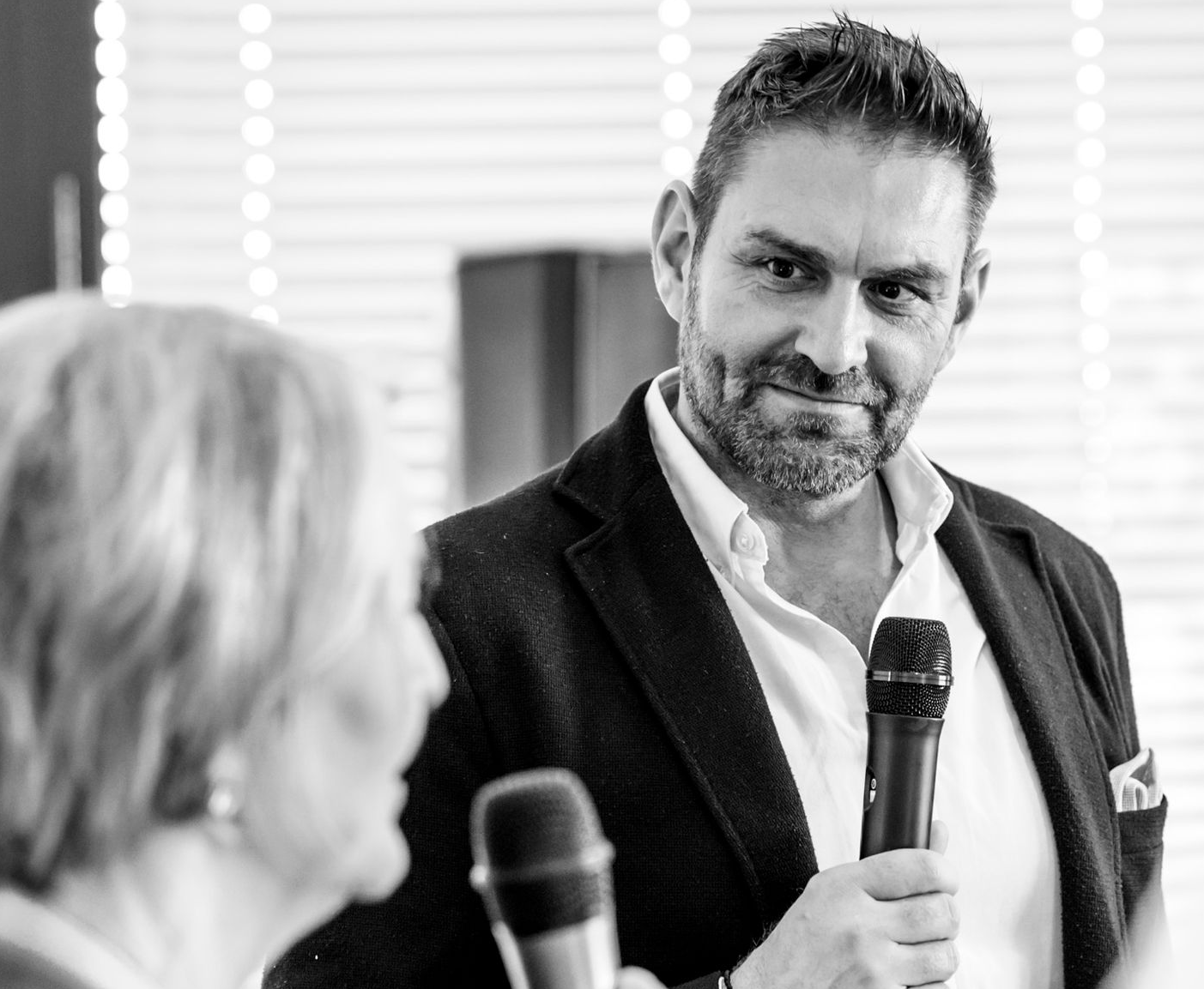 Mann hält ein Mikrofon, lächelt und blickt zu jemandem außerhalb des Bildausschnitts; Innenraumaufnahme, Schwarzweiß.