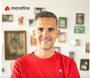 Lächelnder Mann im roten Pullover vor einer Wand mit gerahmten Fotos. Morefire-Logo oben links.