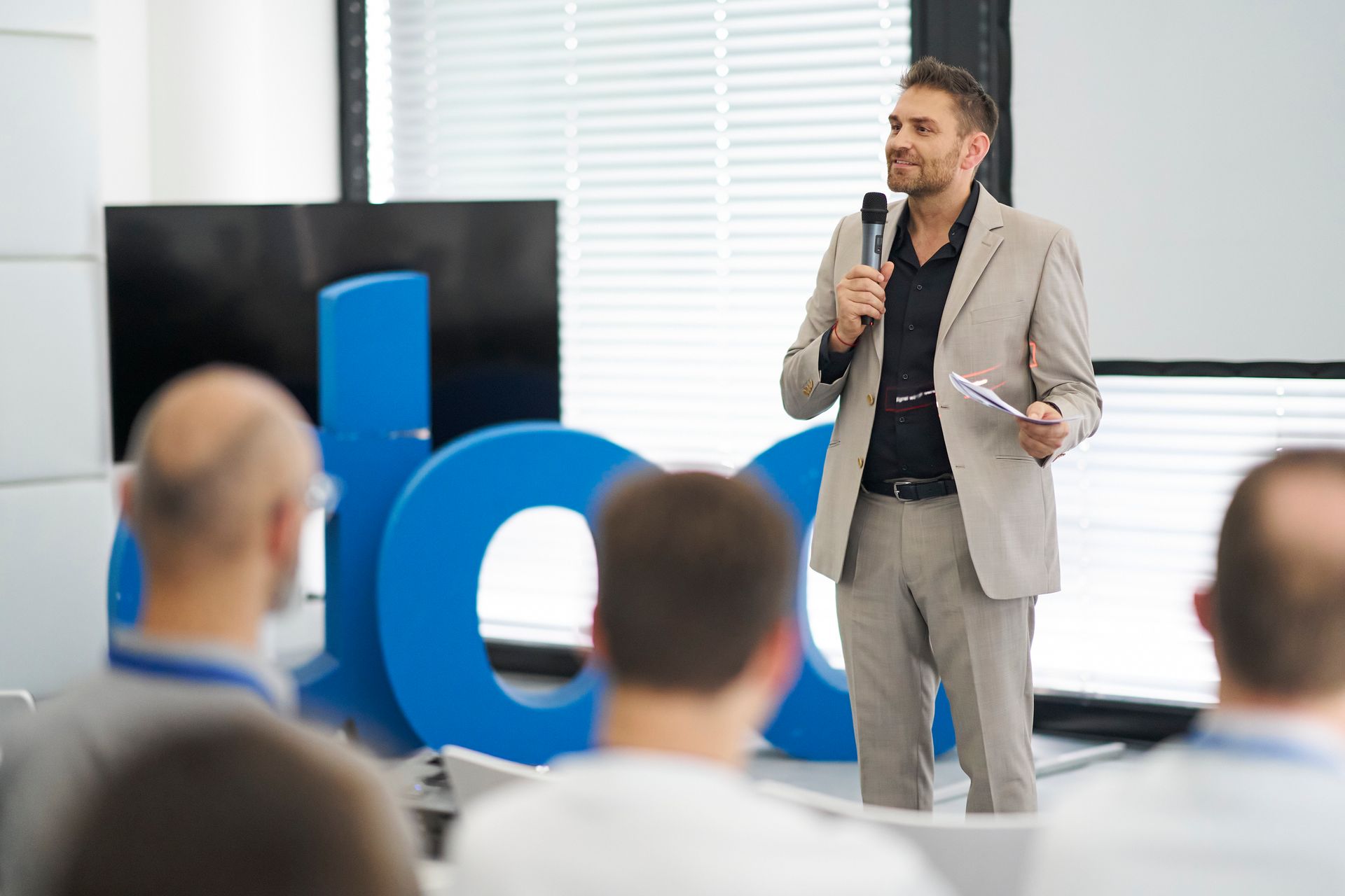 Ein Mann im Anzug spricht auf einer Konferenz in ein Mikrofon; im Hintergrund ist ein blaues Logo zu sehen, das Publikum sitzt.