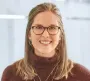 Lächelnde Frau mit Brille und dunklem Pullover in einem hellen Büro.