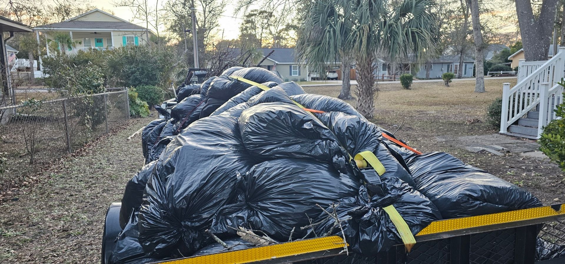 A trailer filled with many black trash bags, secured with yellow straps, parked in a residential neighborhood.