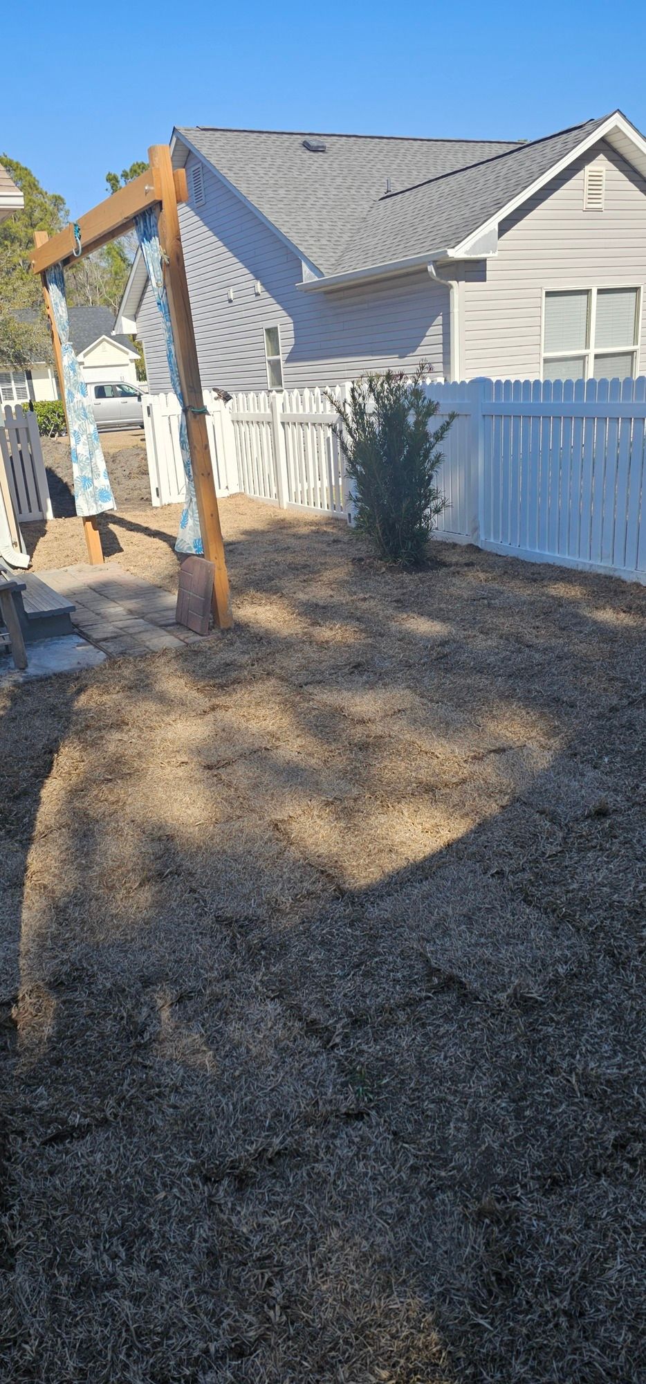 A wooden swing frame stands on a gravel backyard next to a white fence, with a house visible in the background.