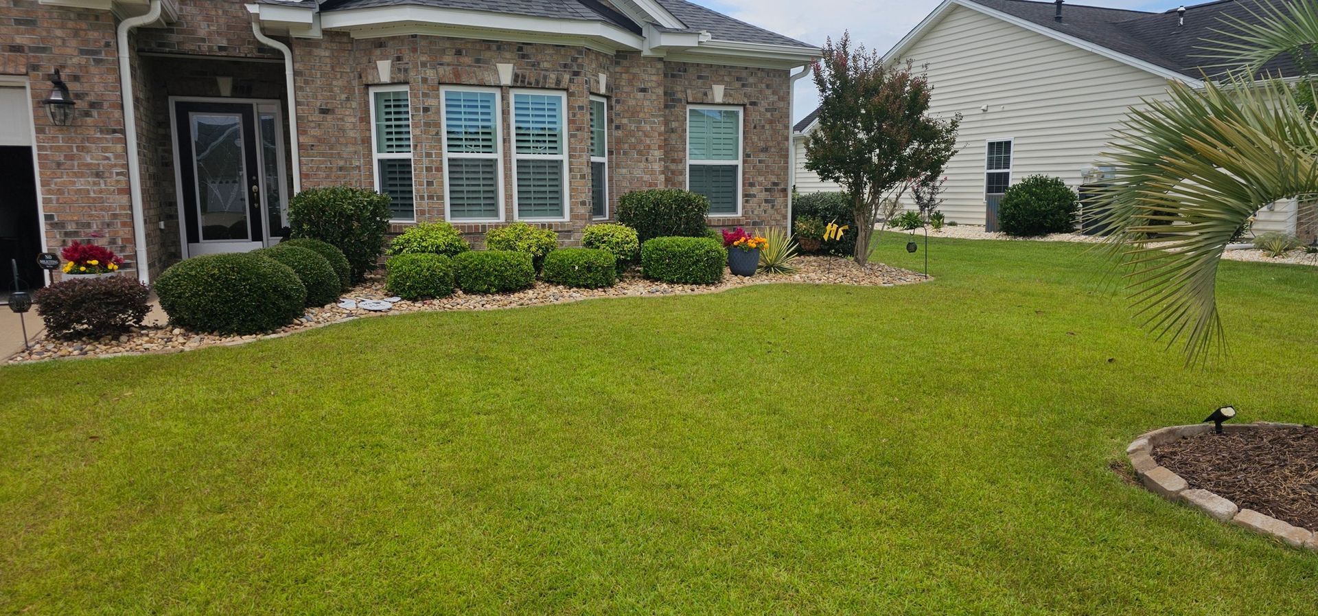 A tan brick house with a green lawn and manicured shrubbery under a cloudy sky.
