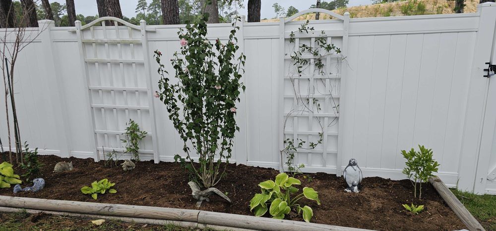 A backyard garden bed features fresh brown mulch, small shrubs, hostas, and two white decorative trellises against a fence.
