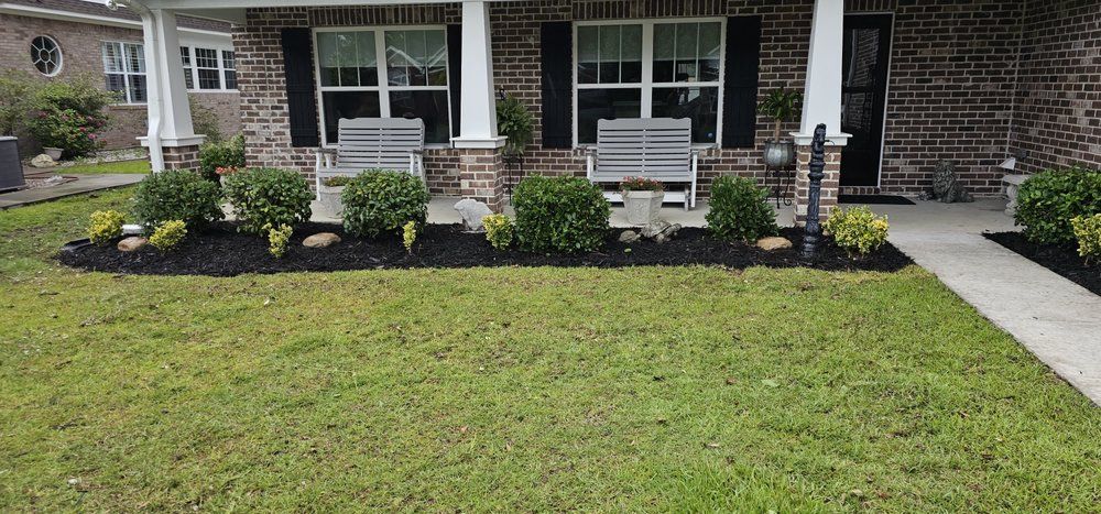 A brick house exterior with a front porch, grey benches, and fresh dark mulch beds featuring green shrubs.