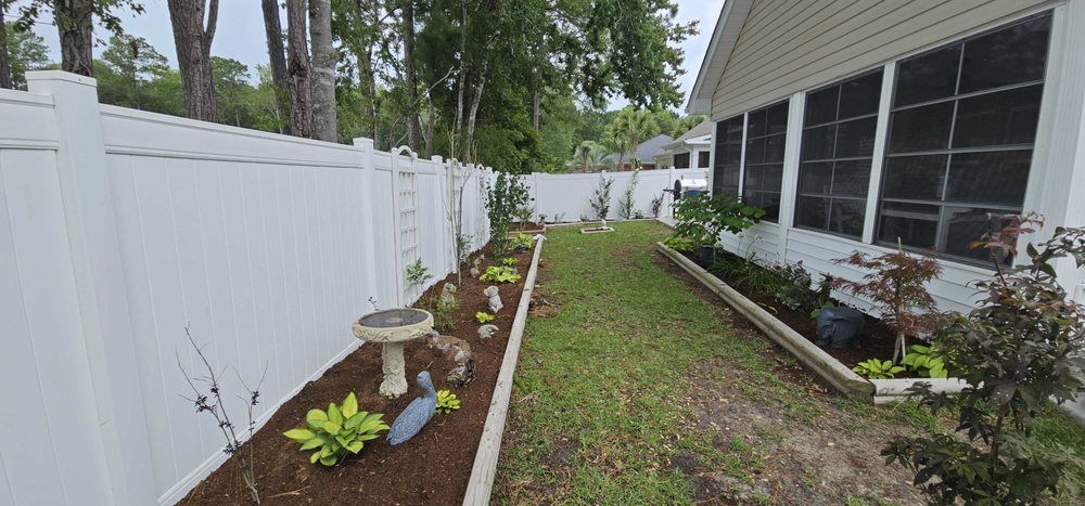 A narrow backyard garden with white vinyl fencing, a bird bath, and planted beds flanking a grassy path beside a sunroom.