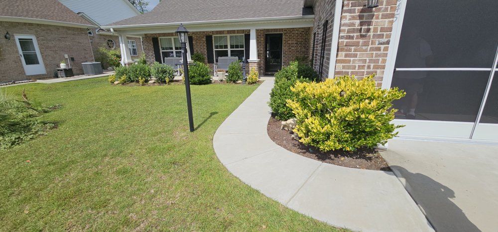 A curved concrete sidewalk leads to a brick home's front entrance, flanked by green grass and a bright yellow bush.