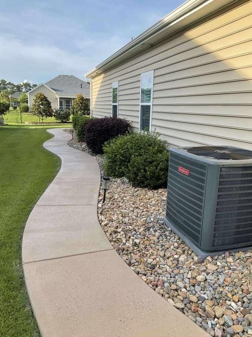 A curved concrete sidewalk leads along the side of a house with tan siding, landscaping bushes, and an outdoor AC unit.