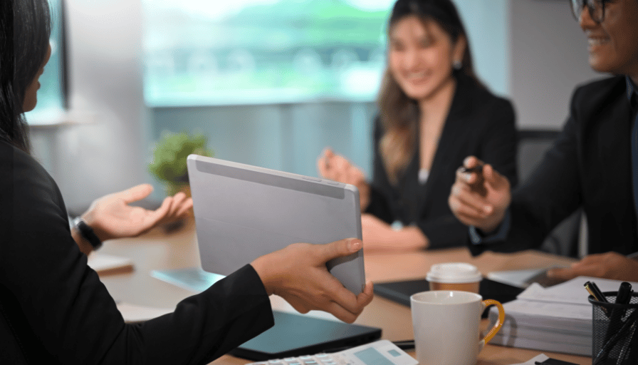 Business meeting with three people around a table, discussing documents and laptops in a bright office.