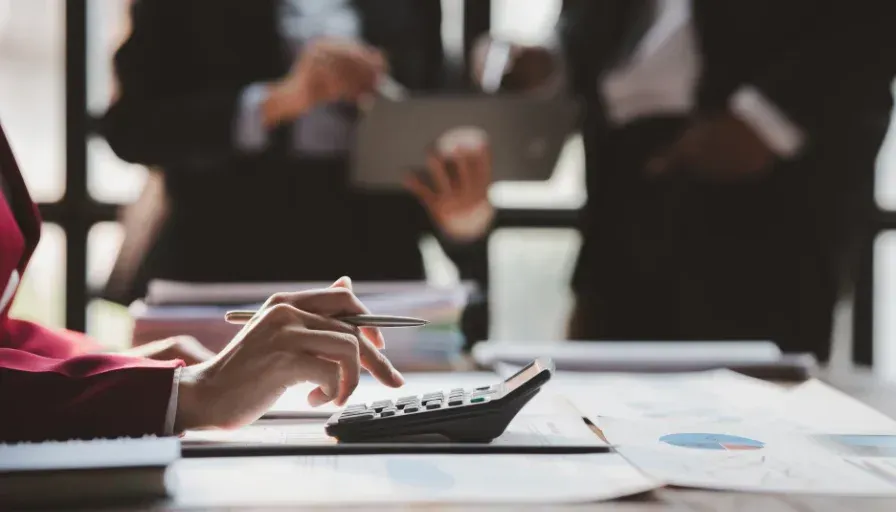 Person using a calculator at a desk during a meeting, with documents and blurred colleagues in the background