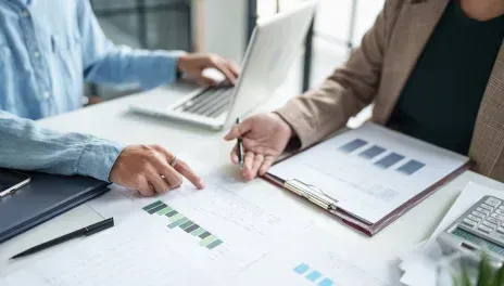 Two people reviewing charts and notes at a bright office desk with laptops and printed reports