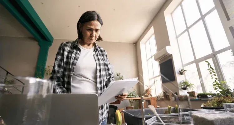 Person reading papers beside a laptop in a bright kitchen with large windows and potted plants