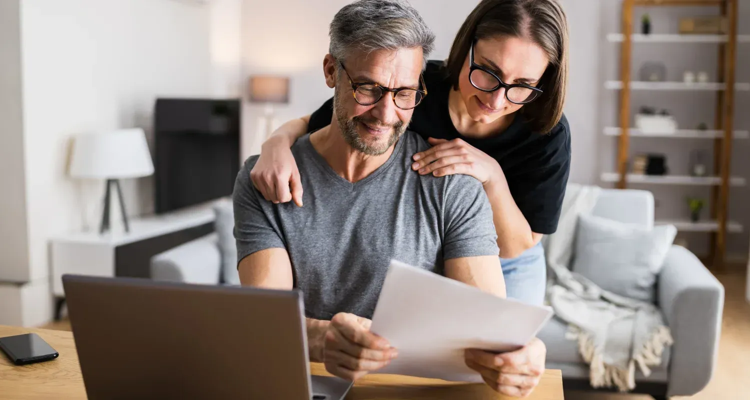 Two people reviewing documents together on a couch with a laptop in a bright living room.