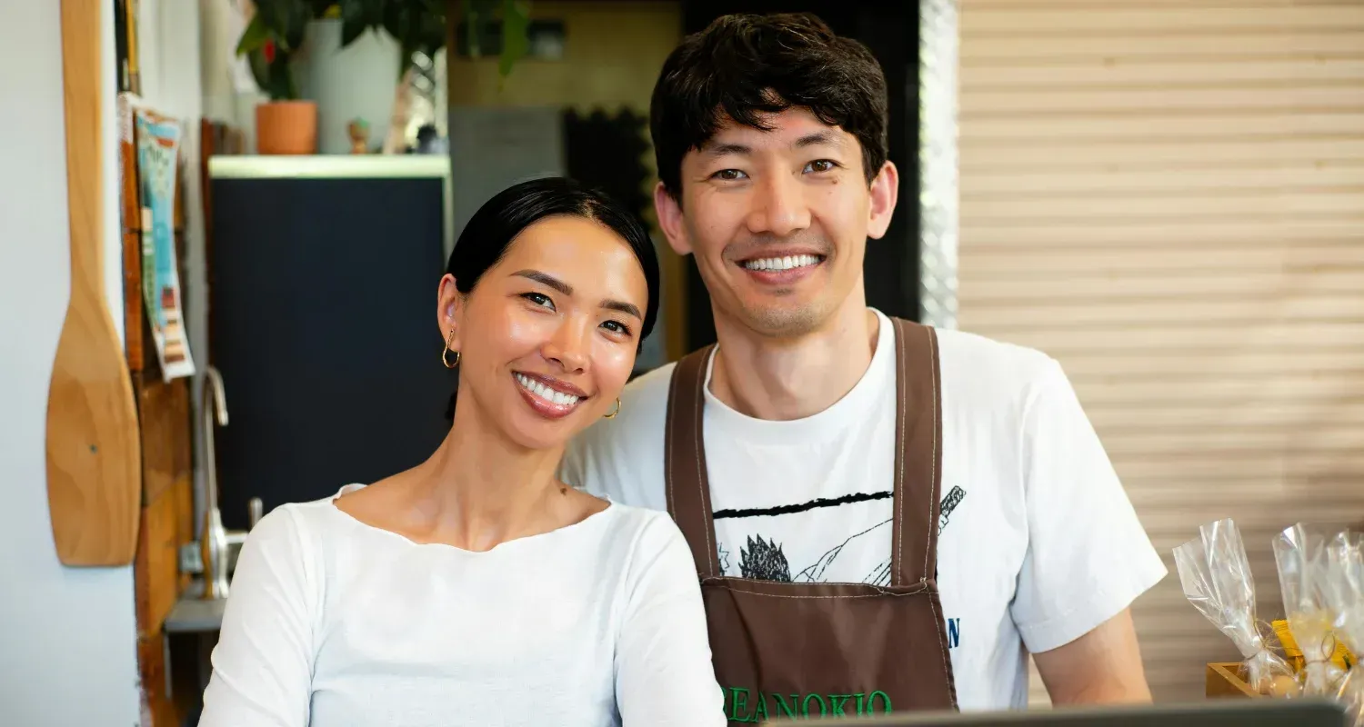 Two smiling people standing behind a counter in a bright café, wearing white shirts and aprons