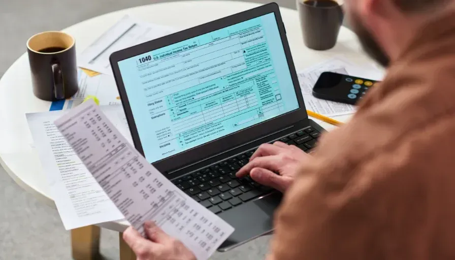 Person reviewing printed invoice beside a laptop displaying a spreadsheet on a desk