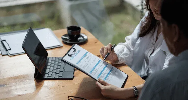 Two people reviewing a clipboard beside a laptop and coffee on a wooden table outdoors