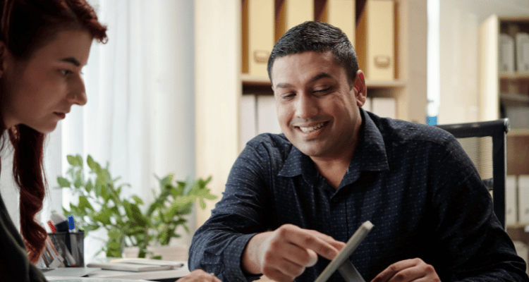 Two people talking at a table, one smiling while holding a tablet in a bright room.