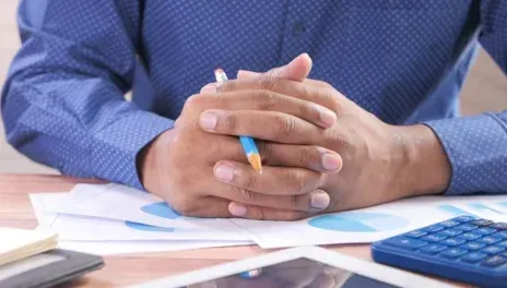 Hands clasped over papers at a desk, with a pencil and calculator nearby