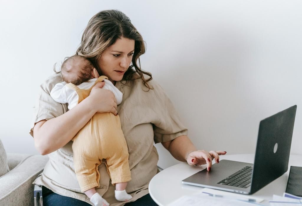 Woman holding a baby while using a laptop at a table in a bright room