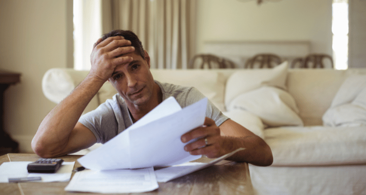 Man sitting at a table, holding papers and rubbing his forehead in a stressed living room.